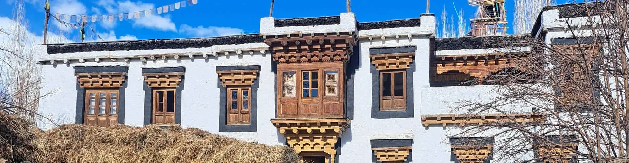 Exterior view of Nimmu House, a traditional Ladakhi-style heritage hotel with whitewashed walls, wooden windows, prayer flags, dry branches, and hay stacks under a blue sky.